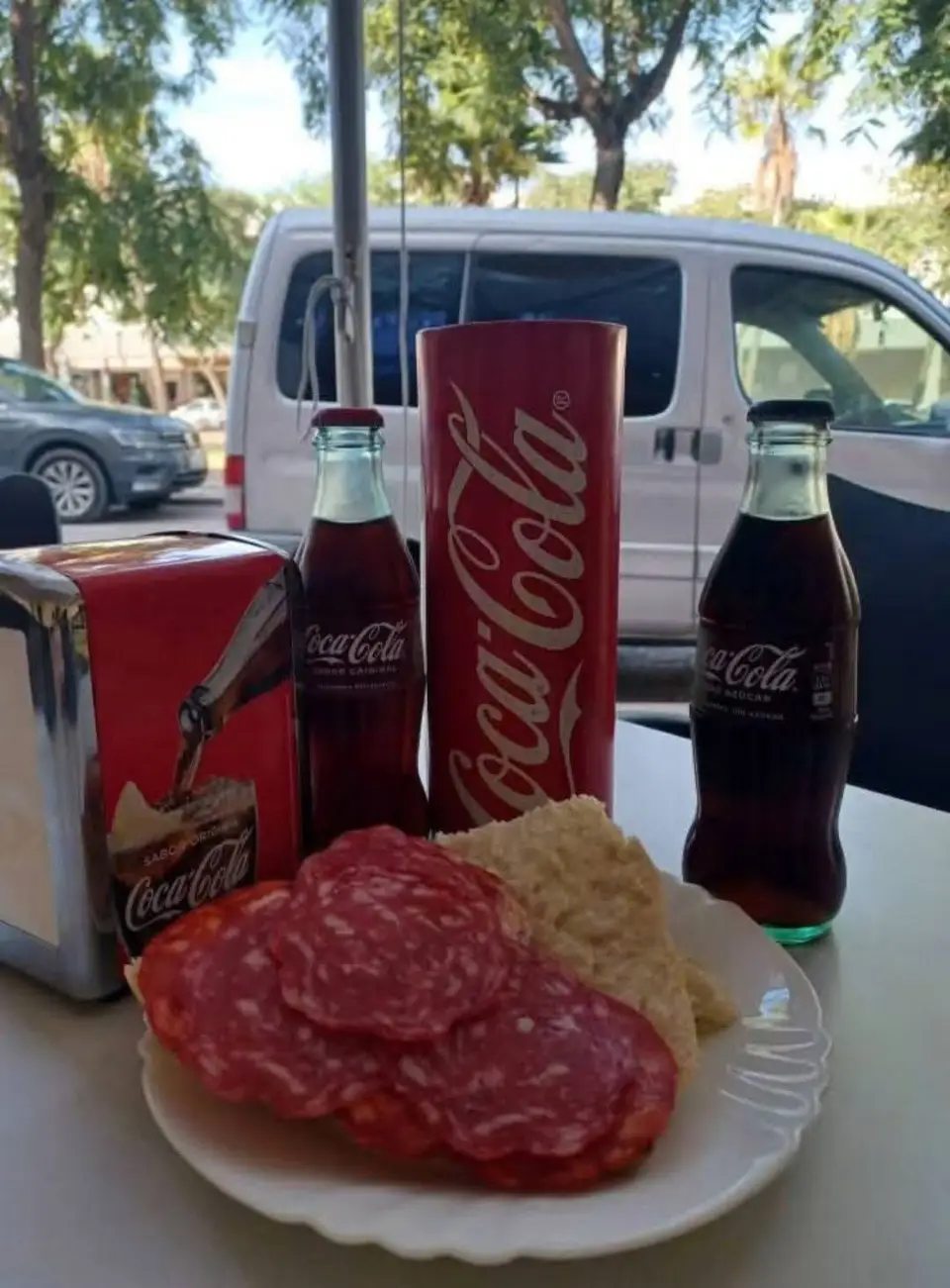 Mesa de la terraza con salchichón, pan y refrescos, con la calle arbolada al fondo